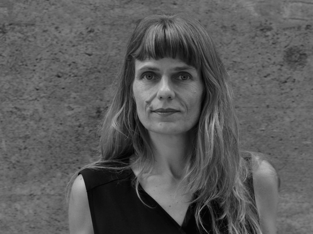Portrait von Katharina Rosenberger. Sie steht vor einer Betonmauer und schaut mit zurückhaltendem Lächeln in die Kamera. 