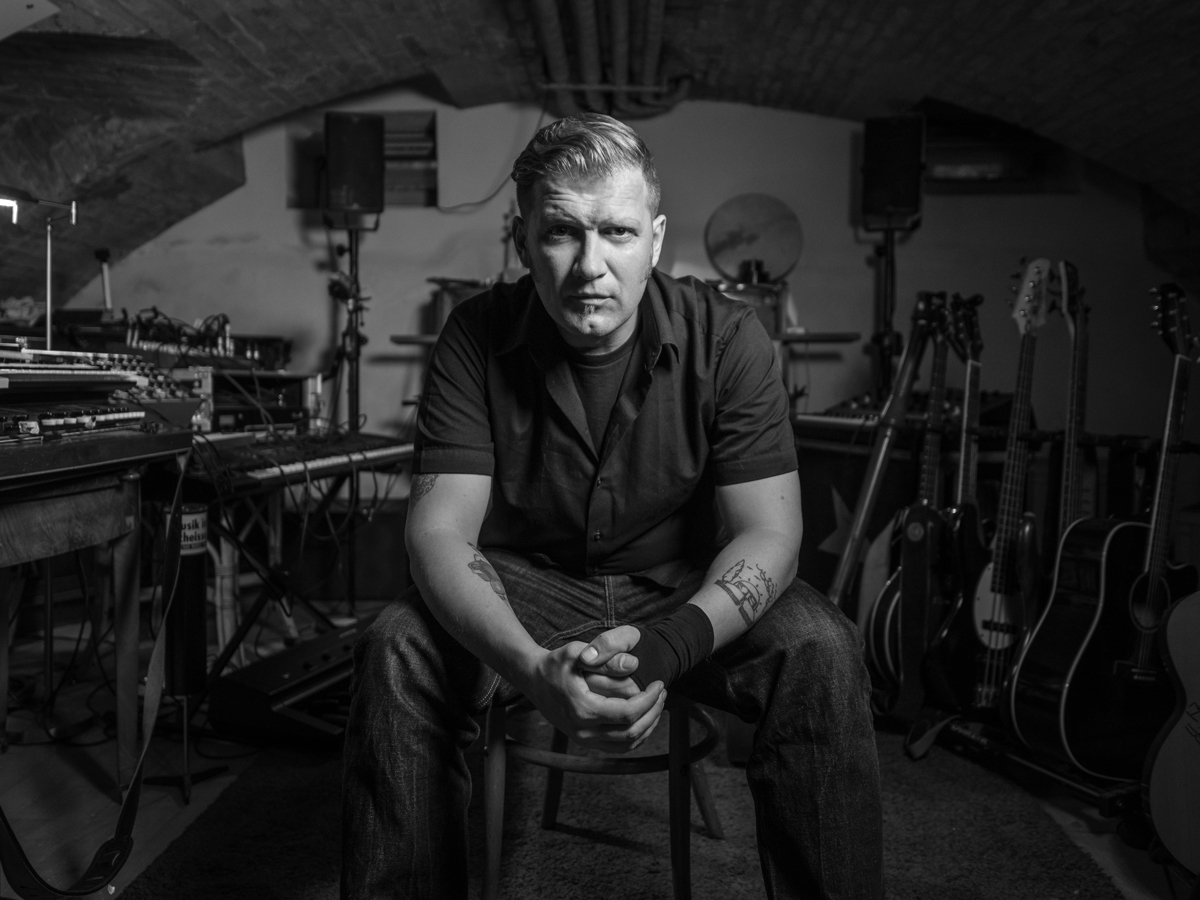 Portrait de Mario Batkociv. Il est assis sur une chaise, le regard directement tourné vers la caméra. A l'arrière-plan se trouve une multitude d'instruments divers.